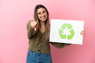 Wall Mural - Young caucasian woman isolated on pink background holding a placard with recycle icon and pointing to the front