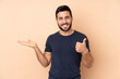 © luismolinero - Caucasian handsome man isolated on beige background holding copyspace imaginary on the palm to insert an ad and with thumbs up