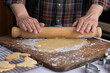 Selective focus angled semi-close-up of the hands and forearms of a person rolling dough on a wooden board with flour dusting,  some undecorated cookies on a wire rack and some cookie cutters.