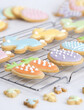 Selective focus angled close-up of pastel coloured homemade Easter cookies on a wire rack against a light background, showing carrot, rabbit, butterfly and egg designs.