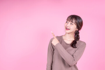 Young attractive Asian girl with Braces smiling happy finger pointing finger blank sign to side, product sales advert banner advertisement, casual wear brown jumper copyspace pink isolated background