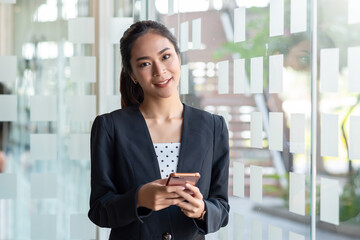 Wall Mural - Asian businesswoman standing smiling holding smartphone. Looking at camera.