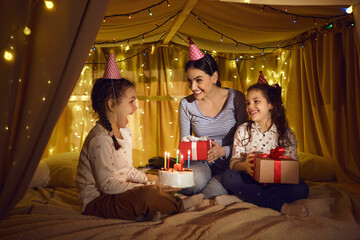 Wall Mural - Family in festive hats celebrates a child's birthday sitting in a cozy tent bed decorated with yellow LED lights. Little girl is going to blow out the candles on the cake. Concept of a creative family