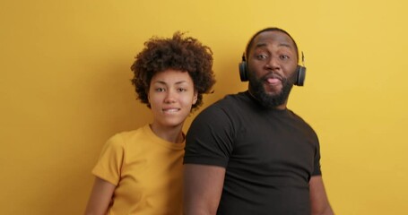 Wall Mural - Positive dark skinned young African American woman and man point directly at camera have fun move energetically enjoy favorite music wear casual clothes isolated over vivid yellow background.