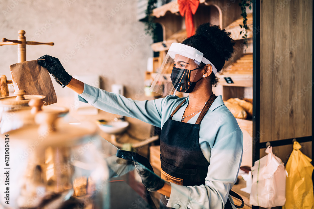African American middle aged female worker with shield and protective mask on face working in bakery. Coronavirus, Covid-19 concept. - obrazy, fototapety, plakaty 