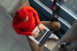 © Yury - Above view of young freelancer man in glasses wear red hat and wool sweater sitting on bench next by window legs crossed, remote online working on laptop, typing on keyboard. Distance job.