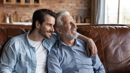 Wall Mural - Smiling older Caucasian father and adult millennial son sit rest on sofa at home look in distance dreaming thinking. Happy mature 60s dad and grownup man kid enjoy family weekend reunion together.