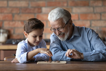 Wall Mural - Smiling senior 60s grandfather sit at table have fun play checkers draughts with small 7s grandson. Happy older granddad teach little smart grandchild, engaged in logical set or game at home together.