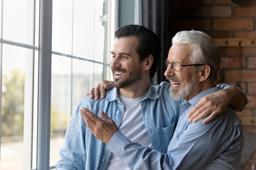 Smiling senior grey-haired Caucasian dad and adult son look in window distance thinking planning together. Happy mature 60s father and grownup man child enjoy family weekend, dream or visualize.