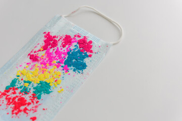 Blue medical face mask, with bright multicolored holi paints, on white background, close-up. Concept of celebrating indian spring festival of colors during covid-19 pandemic. Copy space.