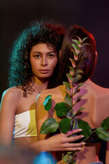 Wall Mural - Creative shot of two brunette women with different hair types posing together, holding green plant, standing in neon light isolated over black background