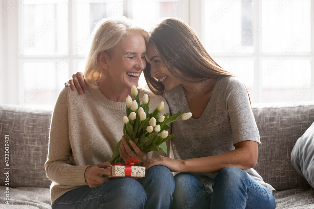 Loving young adult female child congratulate excited elderly mother with birthday anniversary at home. Smiling caring grownup millennial daughter present gift flowers to old mom on women s day. - obrazy, fototapety, plakaty 
