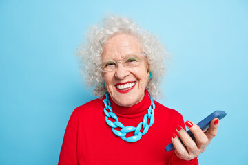 Using modern technologies among all ages. Mature positive grey haired woman with bright makeup dressed in red jumper with necklace uses smartphone waits for call smiles positively at camera.