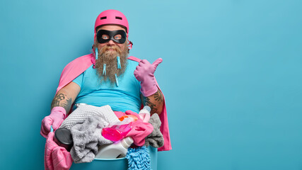 Busy bearded man with thick beard does laundry at home wears eyemask helmet and cloak points thumb away on copy space isolated over blue background holds basin full of dirty linen detergents