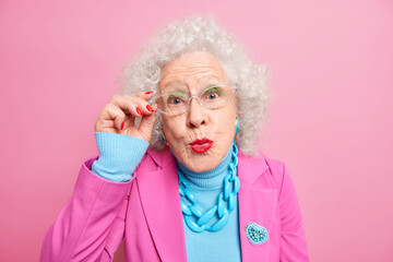 Portrait of senior European woman with curly grey hair keeps hand on rim of spectacles keeps lips rounded dressed in fashionable clothes isolated over pink studio background wants to kiss someone