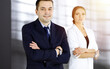 © cameravit - Portrait of a friendly middle aged businessman in a dark blue suit, standing with crossed arms together with a colleague in a sunny modern office. Modern business concept