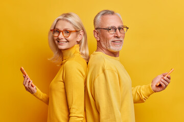 Wall Mural - Photo of cheerful senior woman and man stand in profile with modern gadgets hold smartphone have technology addiction wear casual clothes isolated over yellow background. Pensioners use devices