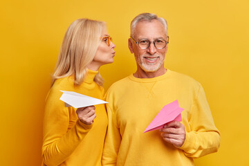 Wall Mural - Pleased beatded grey haired man gets kiss from wife pose next to each other hold handmade paper planes isolated over yellow background. Elderly couple feel love in their old age. Relationship