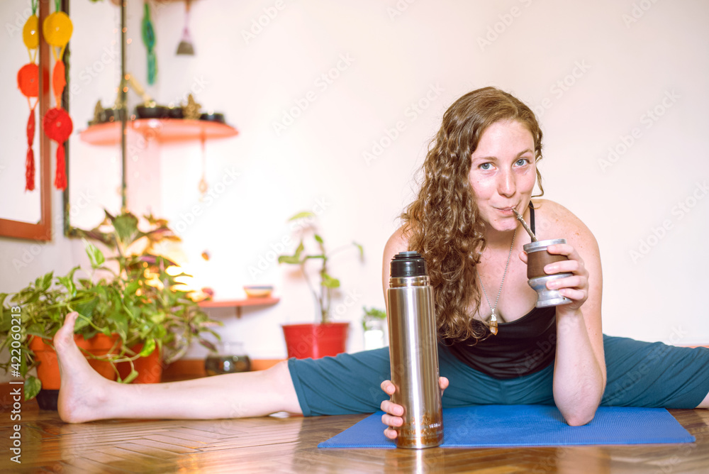 Friendly argentinian yogini teacher drinking traditional yerba mate with bombilla stick. Happy and healthy young girl on fitness mat with spread legs wide open - obrazy, fototapety, plakaty 