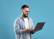 © Prostock-studio - Confident young guy in glasses using laptop pc for online work or communication on blue studio background