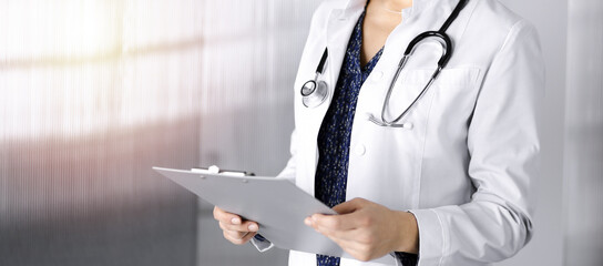 Cheerful intelligent woman-doctor is holding a clipboard in her hands, while standing in a sunny clinic office. Portrait of friendly physician woman with a stethoscope. Perfect medical service in a