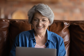 Age and new tech. Excited mature retired hispanic woman in eyewear looking at laptop screen surf internet read good news in email. Happy old aged latina lady sit on sofa use pc hold video call in app