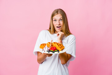 Wall Mural - Young russian woman eating a waffle isolated praying for luck, amazed and opening mouth looking to front.