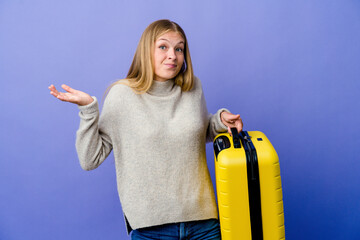 Young russian woman holding suitcase to travel doubting and shrugging shoulders in questioning gesture.