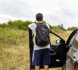 Brunette man with a backpack and cap wearing a gray t-shirt and slippers in front of the open door of a car on a path next to a secluded beach looking up at the sky on a cloudy, rainy day.