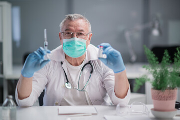 An elderly, gray-haired doctor in a spacious, bright office holds a vaccine against the covid - 19 virus strain and a syringe. Demonstrates a new drug to the camera.