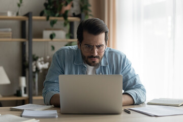 Wall Mural - Close up puzzled focused businessman wearing glasses looking at laptop screen, employee or freelancer working on difficult research project online, reading bad news, analyzing financial report