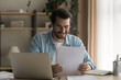 © fizkes - Close up smiling businessman wearing glasses reading good news in paper letter, sitting at work desk with laptop, happy satisfied man holding documents, notification, job promotion, loan approval