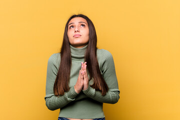 Young Indian woman isolated on yellow background holding hands in pray near mouth, feels confident.