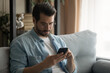 © fizkes - Close up serious man wearing glasses using smartphone, sitting on couch at home, focused young male looking at phone screen, reading news, browsing online banking service, searching information