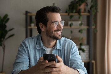 Wall Mural - Close up smiling dreamy man wearing glasses distracted from phone, looking to aside, holding smartphone, visualizing good future, waiting for call or message, enjoying leisure time with gadget