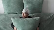 Conspiracy Theorist Man Wearing Tin Foil Hat Hiding Behind Pillow While Sitting On Couch At Home. Paranoid Guy Belief That Aluminium Cap Shield Brain From Mind Control Or Electromagnetic Fields.