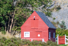 A Vibrant Old Red Wooden Clapboard Building With White Trim. The Old Shed Has A White Circle On The Wooden Door. The Vintage Storage Building Is Among Lush Trees On A Hillside. There's A White Fence.
