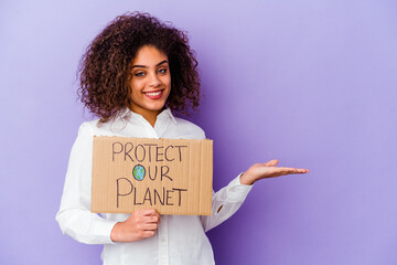 Wall Mural - Young African American woman holding a girl power placard isolated on purple background showing a copy space on a palm and holding another hand on waist.