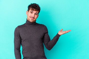 Wall Mural - Young caucasian man isolated on blue background showing a copy space on a palm and holding another hand on waist.