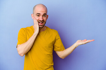 Young caucasian bald man isolated on purple background holds copy space on a palm, keep hand over cheek. Amazed and delighted.