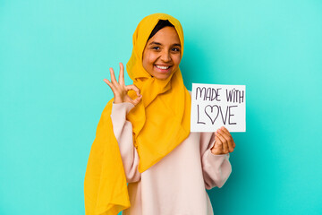 Young muslim woman holding a made with love placard isolated on blue background cheerful and confident showing ok gesture.