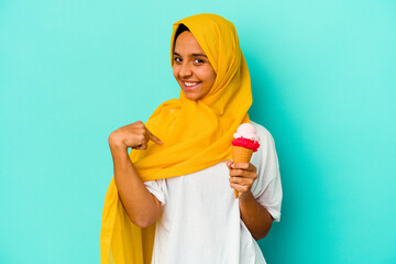 Wall Mural - Young muslim woman eating an ice cream isolated on blue background person pointing by hand to a shirt copy space, proud and confident