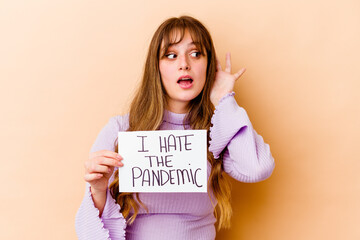 Wall Mural - Young caucasian woman holding a I hate the pandemic placard isolated trying to listening a gossip.