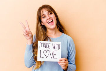 Wall Mural - Young caucasian woman holding a made with love placard isolated joyful and carefree showing a peace symbol with fingers.