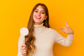 Wall Mural - Young caucasian woman holding a lollipop isolated on yellow background person pointing by hand to a shirt copy space, proud and confident