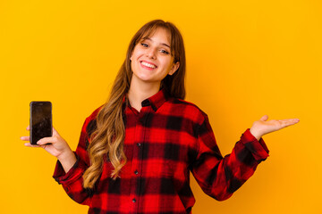 Wall Mural - Young caucasian woman holding phone isolated on yellow background showing a copy space on a palm and holding another hand on waist.