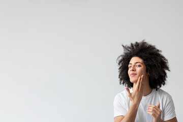 Man skincare. Facial treatment. Male grooming. Satisfied handsome guy with long brown curly hair applying lotion after shaving on smooth face skin isolated on neutral empty space advertising banner.