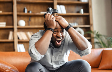 Front view portrait of young African American guy sitting on the couch in the living room, holding head in hands and joystick, screaming because lost video game match, male addicted gamer feeling mad