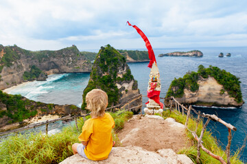 Wall Mural - Happy boy have fun on summer beach holiday. Little child relax at rock cliff above sea. Looking at beautiful view of Atuh beach. Healthy family lifestyle, summer travel on tropical island.