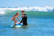 © Tropical studio - Little surf girl - young surfer learn to ride on surfboard with instructor at surfing school. Active family lifestyle, kids water sport lessons, swimming activity in summer camp. Vacation with child.